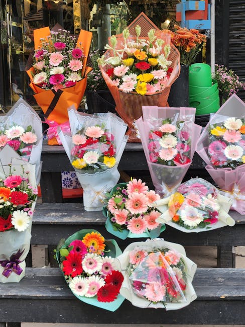 A display of various floral bouquets arranged on wooden steps outside a florist shop. The bouquets feature a mix of fresh flowers including pink, white, yellow, and red daisies, chrysanthemums, and ca