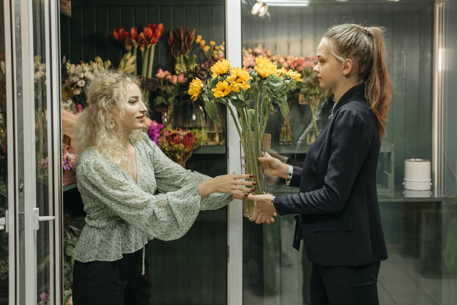 A florist with curly blonde hair, wearing a light green blouse with a ruffled hem, is handing a bouquet of yellow sunflowers to a female customer with long brown hair tied back in a high ponytail, dre