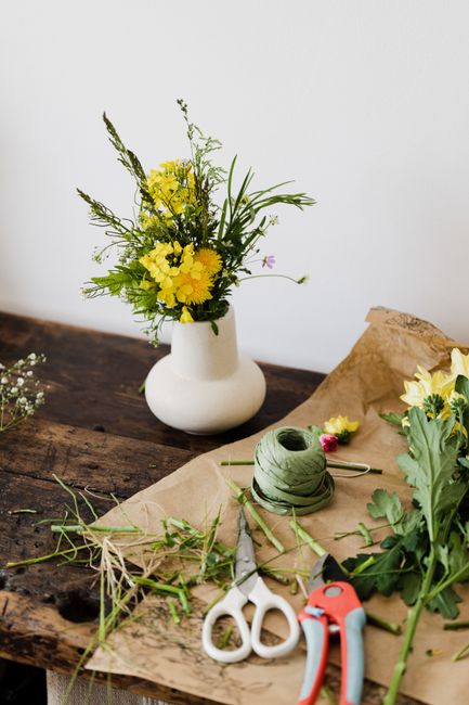 A fresh floral arrangement in a white, round ceramic vase featuring yellow flowers, including daffodils and other blooms, complemented by green foliage and tall, slender stems, is placed on a rustic w