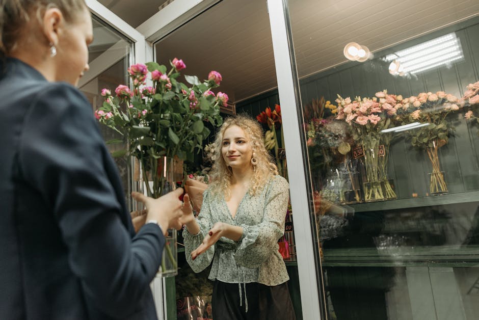 A woman with curly blonde hair, wearing a light green floral blouse, is receiving a bouquet of fresh pink roses with green leaves from another person inside a flower shop. The bouquet is wrapped in br