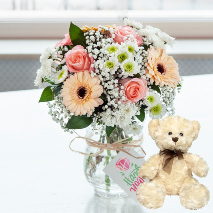 Set against a luminous, softly blurred background that recalls the gentle daylight filtering into a Little Ilford living room, a pastel bouquet and a teddy bear create a tender anniversary tableau. The arrangement is presented in a clear glass jar-style vase placed on a flawless white surface that lightly reflects the colours above it. Around the neck of the vase, a piece of natural jute twine is tied in a modest bow, from which hangs a small white rectangular tag. On the tag, a simple pink heart graphic crowns flowing green script, hinting that these flowers were arranged with a personal, handmade touch, the way a local florist might prepare a surprise for a couple celebrating off High Street North. The bouquet itself is softly rounded and full, with blush pink roses unfolding in delicate layers at the heart of the design. Interspersed among them are peach gerbera daisies whose petals radiate outward from dark brown centres, adding a warm, slightly playful note. Between the larger blooms, airy sprays of white baby's breath create a cloud-like softness, while clusters of neat white chrysanthemums with fresh lime-tinted centres add brightness and structure. Slender glimpses of green foliage weave through the composition, grounding the pastel palette and enhancing its freshness. To the right of the vase, a small cream-coloured teddy bear sits attentively, its plush fur appearing thick and comforting. Dark brown button eyes, a matching nose, and a narrow brown satin ribbon tied neatly around its neck give the bear a look of quiet loyalty, as if ready to keep a secret or share a cherished memory. This intimate pairing of flowers and bear suggests affection, gratitude, and enduring love, fitting for anniversaries and meaningful occasions celebrated in Little Ilford homes.